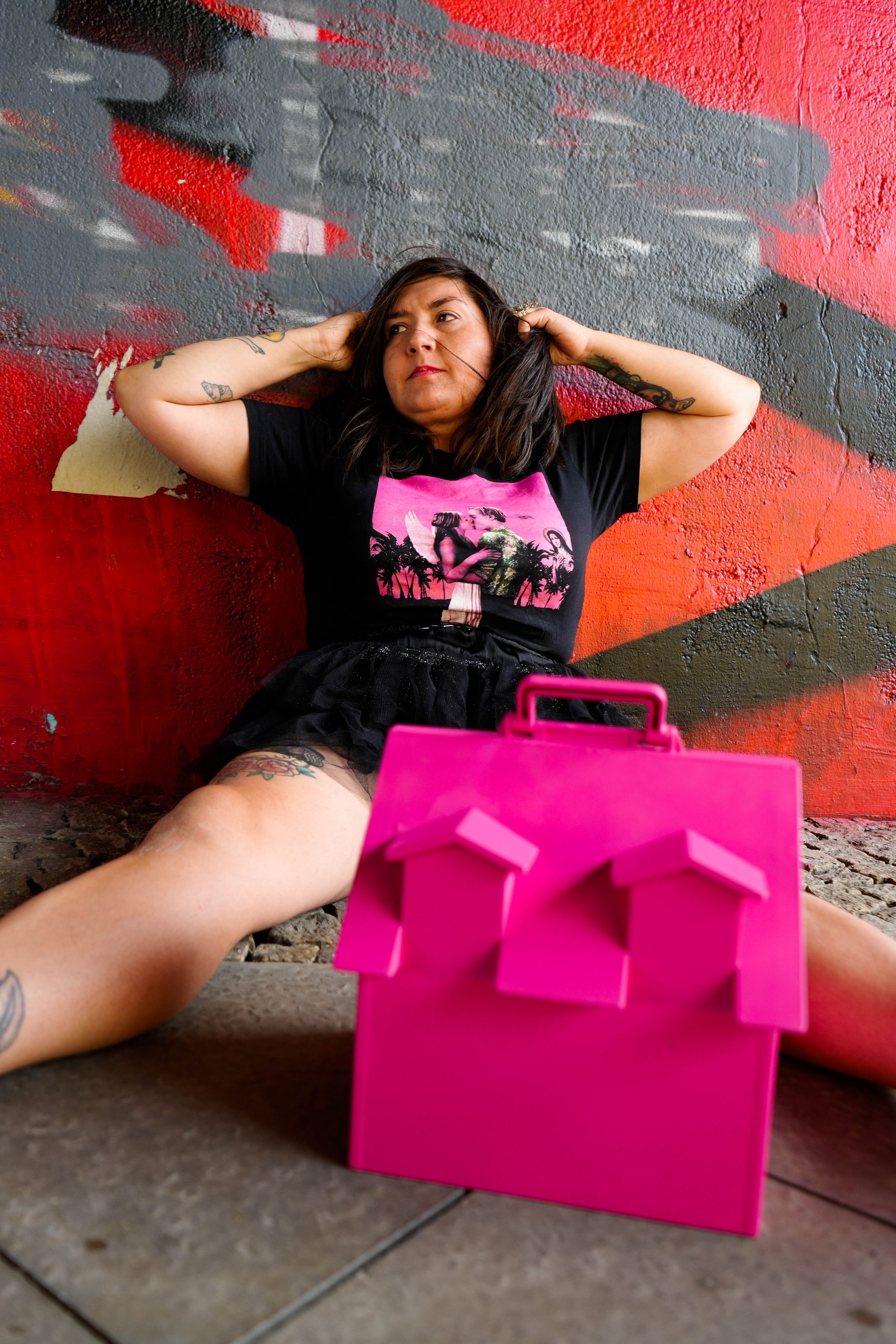 Andrea in a black tulle skirt with her hands in her hair, sitting against a red graffiti wall behind a pink house-shaped bag